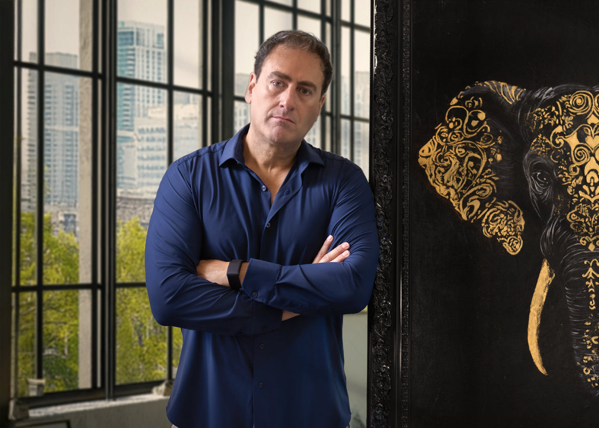 Man in a blue shirt standing next to an ornate black and gold painting of an elephant with a cityscape in the background.